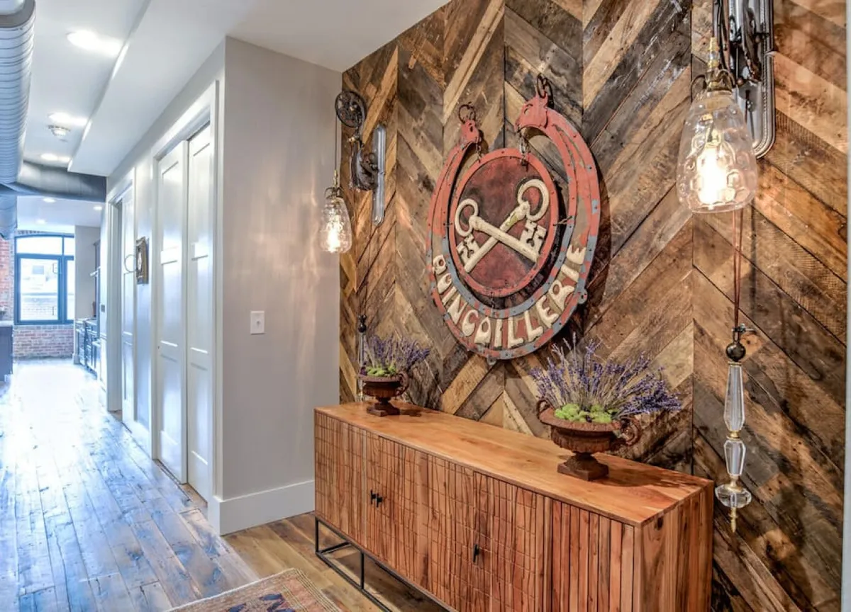 An inviting entryway featuring a wooden herringbone accent wall, a vintage sign, a wooden console table adorned with decorative plants, and hanging light fixtures. The space exudes warmth and offers a glimpse of the home's unique personality right as you step in.