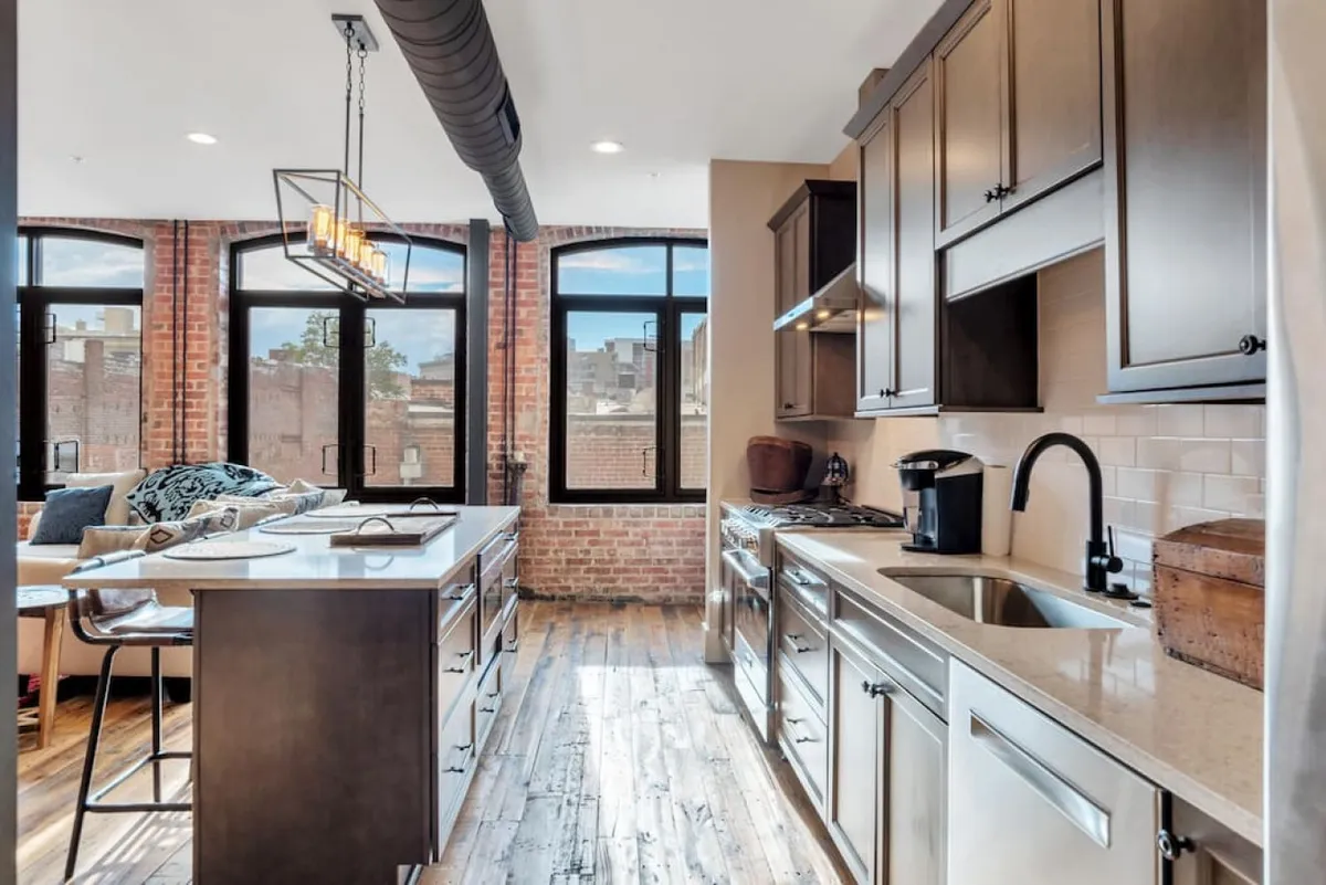 The scene showcases a warm and spacious area with large windows, a kitchen island, and exposed brick walls, giving the room a blend of modern comfort and historical charm. The abundance of natural light and the visible kitchen and dining zone make it ideal for hosting meals and social gatherings.