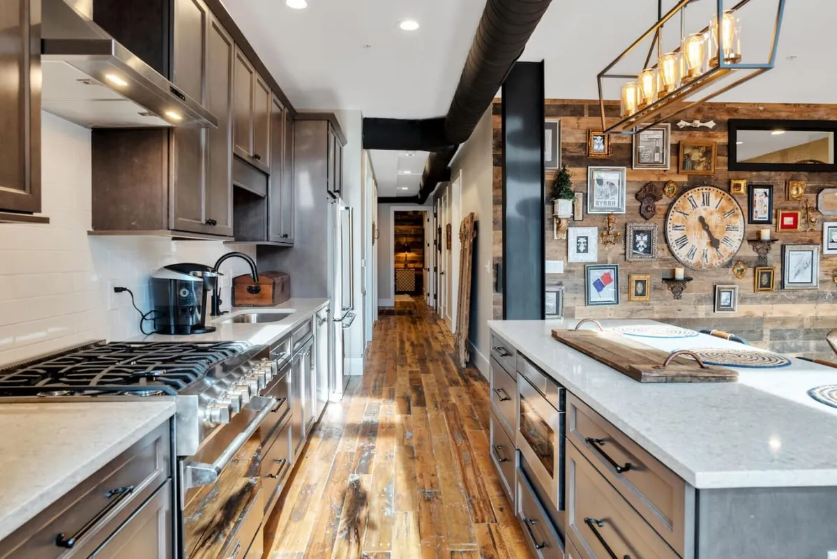 A modern kitchen with stainless steel appliances, a spacious island, a textured brick wall, various framed pictures, and a striking modern light fixture. The scene is designed for both cooking and entertaining, with ample space for preparing meals and hosting gatherings.