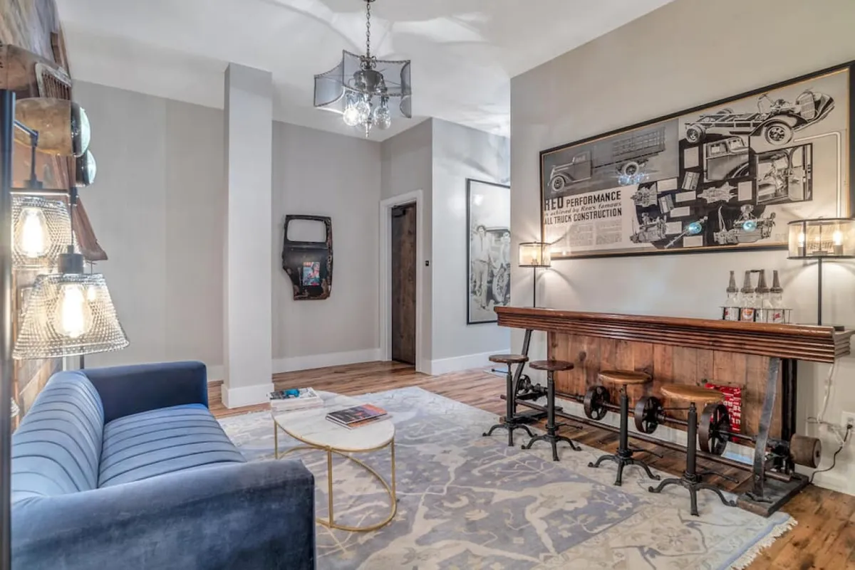 A stylish interior featuring a blue sofa, a wooden bar, unique artwork on the wall, and decorative lighting fixtures. The layout suggests an open, inviting space connected to the living area, ideal for relaxing or informal meetings.