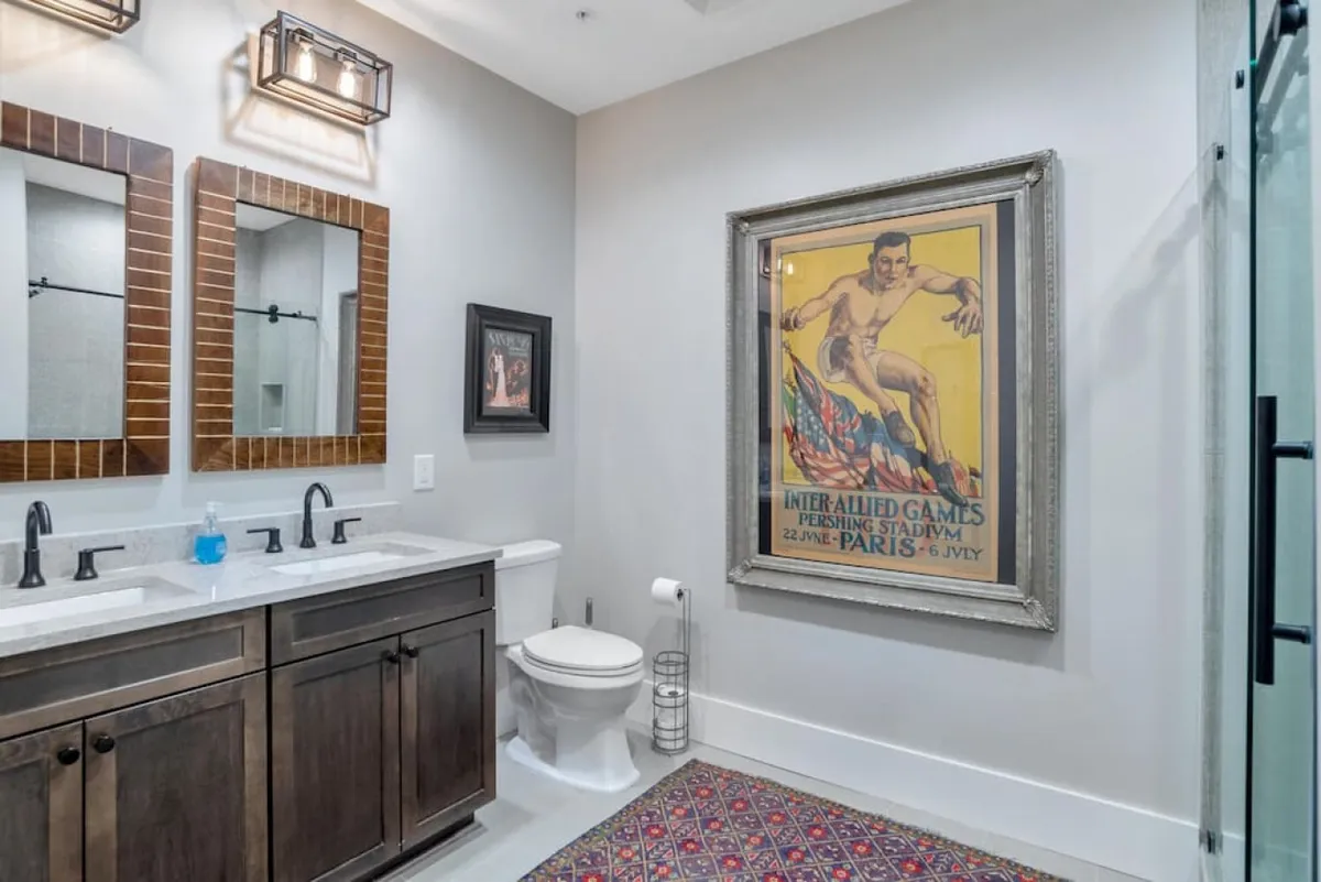 A sophisticated ensuite bathroom with double sinks set in dark cabinetry, a framed vintage poster, and an intricate rug. The mood is both stylish and comfortable, ideal for unwinding or morning routines.