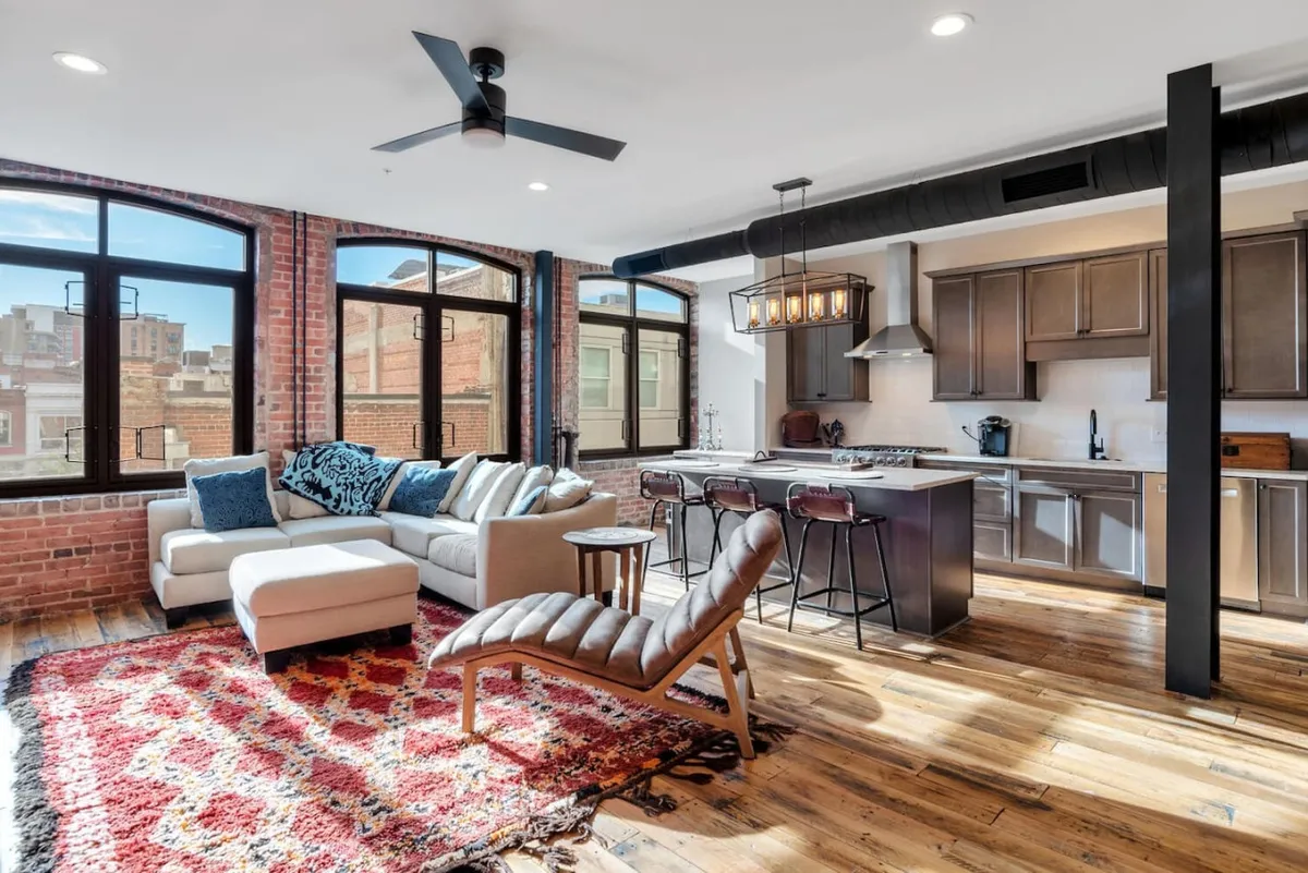 This image captures a cozy and inviting open-concept space featuring a comfortable sofa, a stylish rug, a kitchen island, and oversized windows that let in abundant natural light. The space is designed for both relaxing and entertaining, with an atmosphere ideal for gatherings and unwinding alike.
