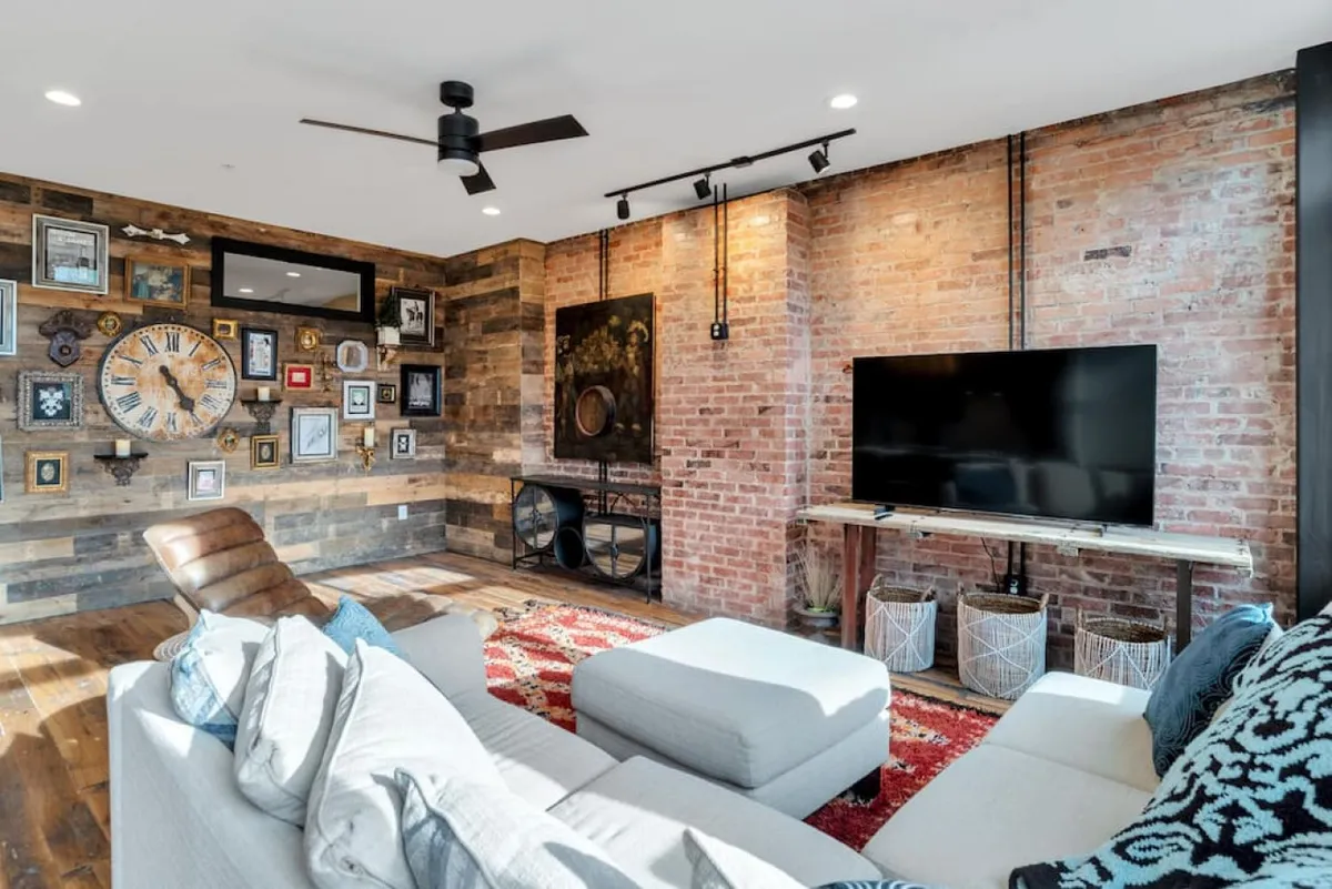 A stylish lounge setting with a prominent brick wall, large clock, TV unit, comfortable sofa, and a modern area rug. Artistic decor and warm lighting create an inviting, conversation-friendly environment ideal for relaxing or entertaining guests.