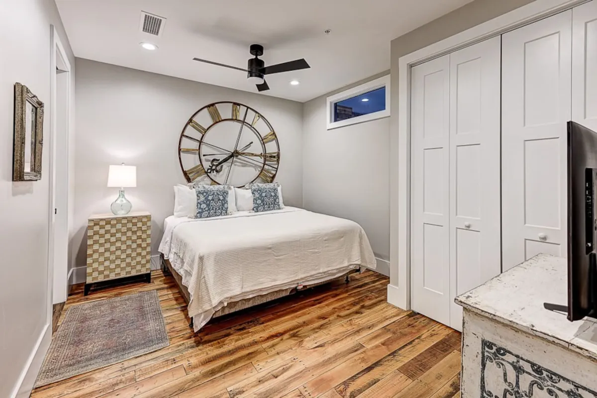 A cozy bedroom scene with a comfortable bed adorned with decorative pillows, a large wall clock, and a nightstand with a lamp. Natural light filters in, suggesting a restful and quiet atmosphere.