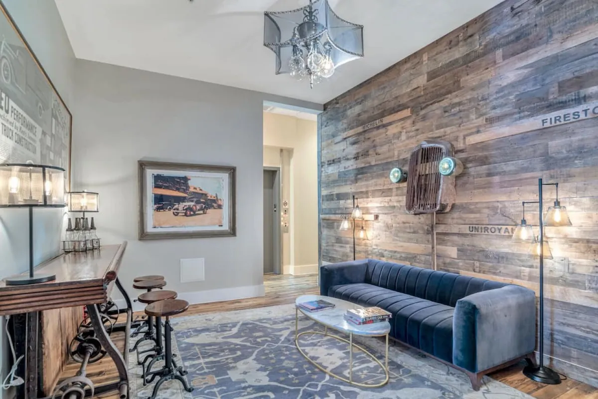 A contemporary living area featuring a blue velvet sofa facing a wooden accent wall, with a modern chandelier overhead and colorful artwork on the wall. There are bar stools in the background, suggesting a space ideal for gathering and socializing.