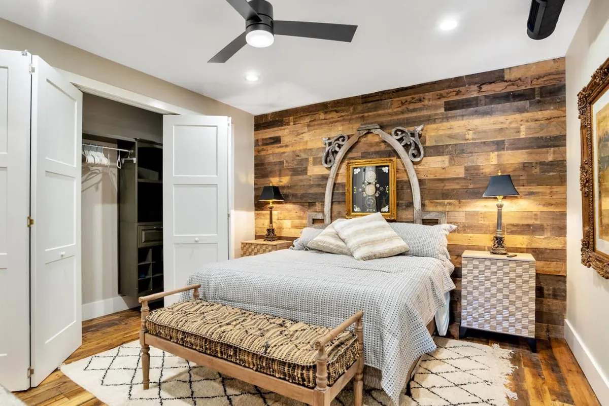 A calming bedroom with a wooden accent wall, textured bedding on the king-size bed, a decorative mirror, and table lamps. The scene exudes serenity and is softly lit, emphasizing relaxation and a peaceful wakeup.