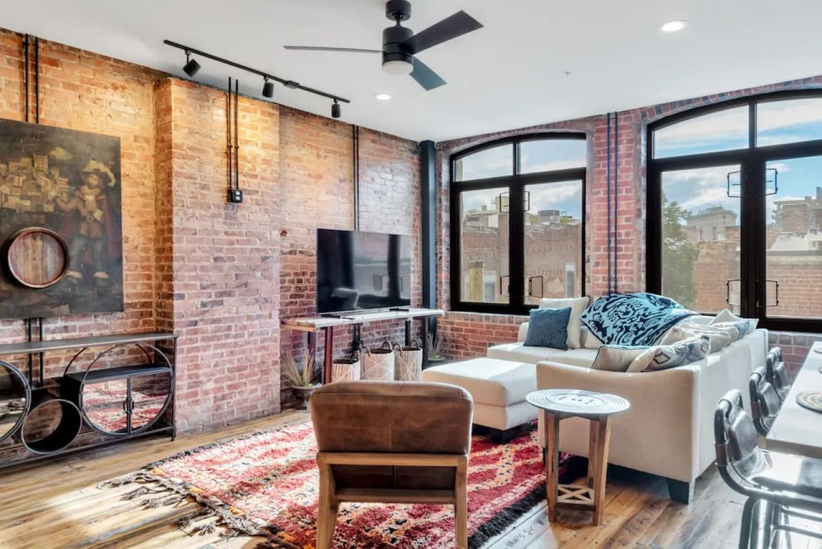 A spacious, bright living room with an exposed brick wall, large windows, a comfortable sofa with decorative pillows, and a TV unit. The room is stylish yet relaxed, ideal for hosting group activities or unwinding after a day of urban adventures.