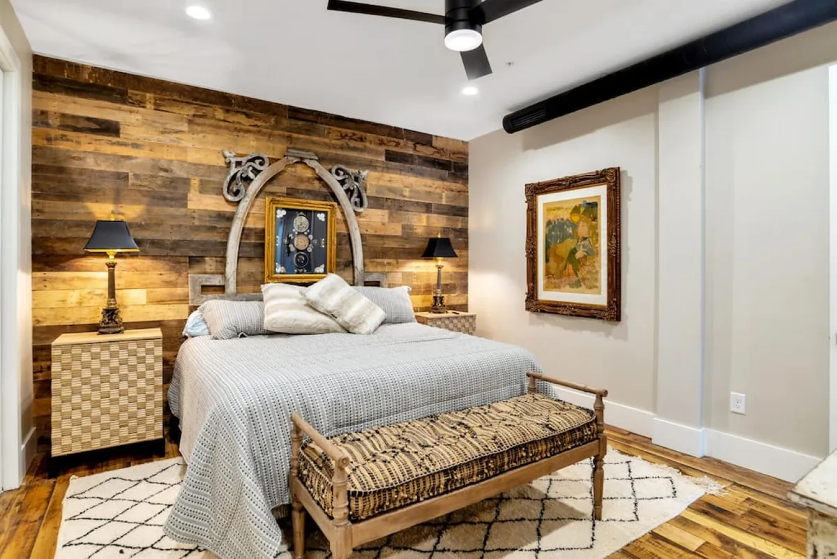 A cozy bedroom with a wooden accent wall, a neatly made bed adorned with decorative pillows, framed artwork, and bedside table lamps. The mood is warm and restful, perfect for unwinding after a night out.