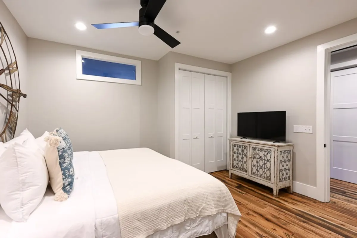 A serene bedroom anchored by a plush king bed with decorative pillows, complemented by a nightstand, television, ceiling fan, and a stylish wall piece. The space is bathed in tranquil lighting, evoking comfort and privacy.